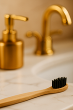 Load image into Gallery viewer, Bamboo toothbrush with black bristles on a bathroom counter with gold faucet and soap dispenser.
