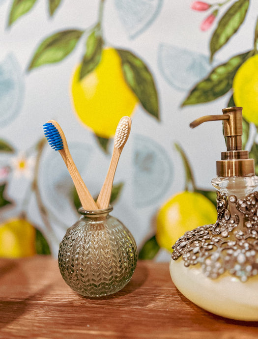 Two bamboo toothbrushes with blue and white bristles in a clear glass holder, placed on a wooden surface against a wall with yellow and green fruit illustrations.